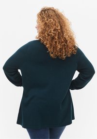 Turkusowy, oversize sweter o miękkiej teksturze, z długimi rękawami i centralnym szwem. Tył ma zaokrąglony dół.