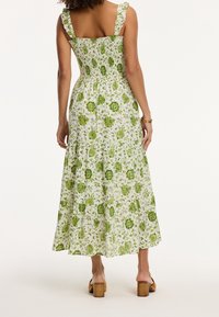Maxikleid mit angepasstem Oberteil, gerafftem Rücken und Rüschenträgern. Weißer Stoff mit grünen Blumenmustern und gestuftem Saum.
