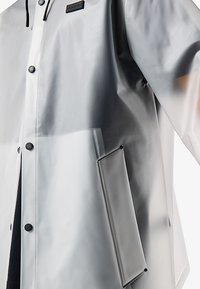 Transluzenter grauer Regenmantel mit schwarzen Druckknöpfen, Seitentasche und "HUNTER"-Logo auf der Brust, getragen über schwarzer Kleidung.