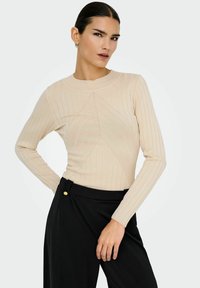 Kvinna med mörkt hår iklädd en beige långärmad ribbad topp och svarta högmidjade byxor mot en ljus bakgrund, poserande med handen på höften.