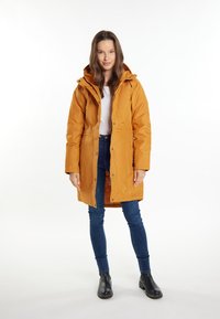 Parka impermeabile gialla con cappuccio, bottoni a pressione e tasche, indossata sopra una maglietta bianca e jeans blu, abbinata a stivaletti neri alla caviglia.
