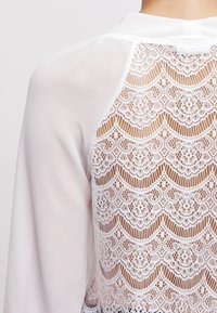 Sheer white blouse with long sleeves and intricate floral lace pattern on the back panel, worn by a person shown from behind.