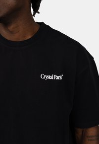 Černé bavlněné tričko s kulatým výstřihem a krátkými rukávy, s bílým logem "Crystal Paris" na levé části hrudi.