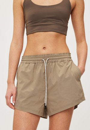 Femme portant un short de sport beige avec cordon de serrage et un brassière de sport marron, visible de la mi-cuisse au haut du torse, debout avec une main dans la poche.