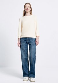 Jasnożółta, miękka bluza z okrągłym dekoltem i rękawami do łokcia; połączona z szerokimi, średnio niebieskimi dżinsami i białymi trampkami.