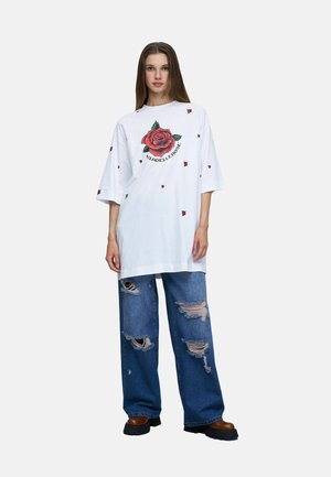 Femme portant une chemise blanche oversize avec un grand motif de rose rouge et un jean bleu déchiré avec des chaussures marron sur un fond blanc.