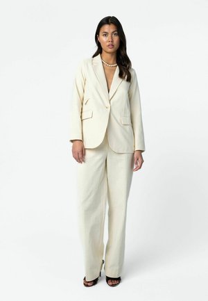 Vrouw met lang donker haar, gekleed in een gebroken wit op maat gemaakte blazer en bijpassende wijde broekspijpen, met zwarte open sandalen en een parelketting.