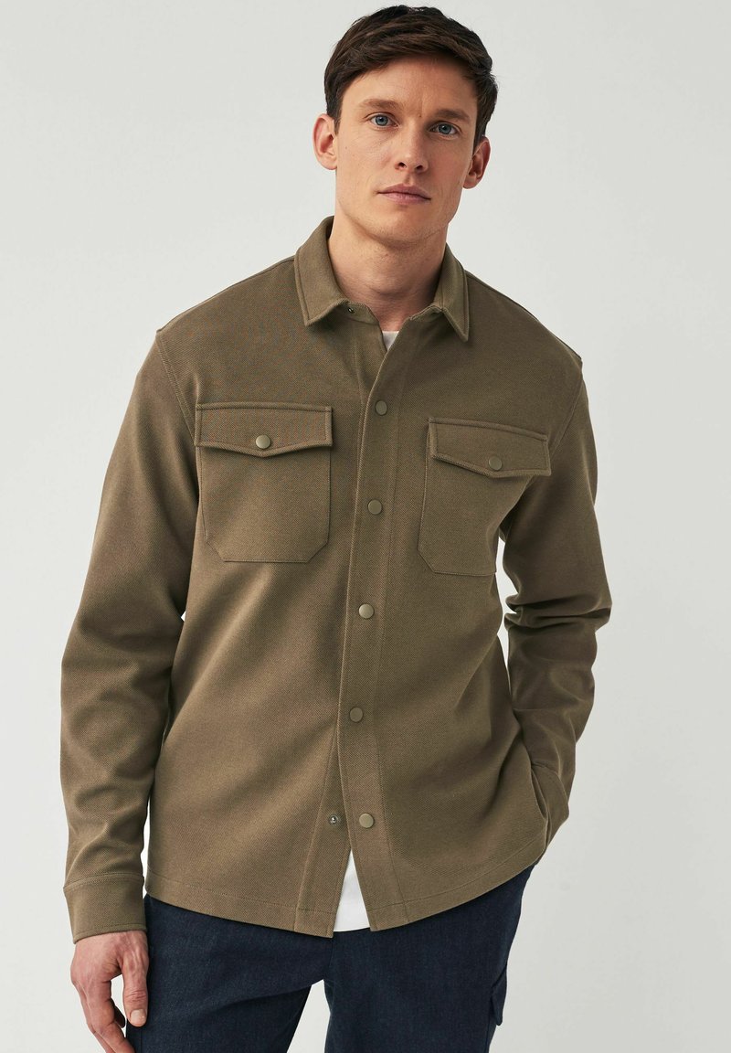 Chaqueta de camisa verde oliva con cierre de botones, dos bolsillos en el pecho y mangas largas. Tejido suave y texturizado, con cierres a presión.