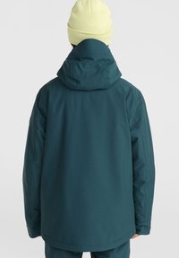 Veste imperméable teal avec capuche, présentant une coupe décontractée et une texture lisse, des coutures visibles et un design uni. Portée avec un bonnet jaune clair.