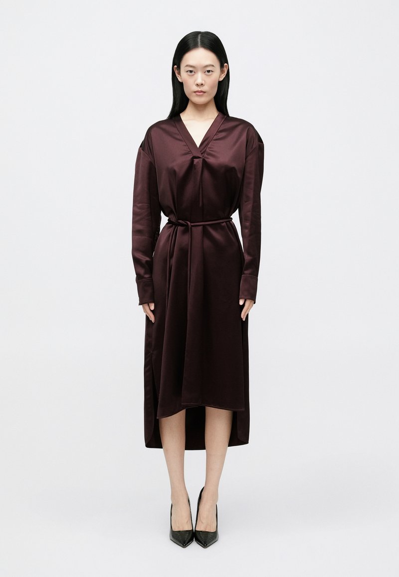 Φόρεμα από σατέν μπορντό με V-λαιμόκοψη και μακρι�ά μανίκια, ζωνάρι στη μέση. Διαθέτει ρέουσα σιλουέτα και πλαϊνές ανοίγματα, συνδυασμένο με μαύρες γόβες.