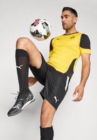 Conjunto atlético negro y amarillo con una camiseta texturizada, pantalones cortos a juego, calcetas negras hasta la rodilla y zapatos de fútbol negros. Balón posicionado sobre el pie.