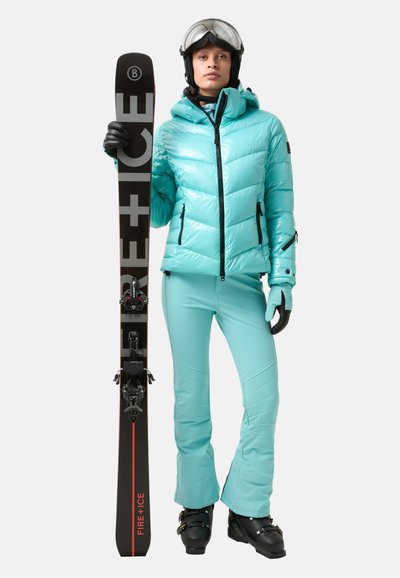 Ski outfit featuring a shiny, light blue puffer jacket and matching ski pants. Black gloves and ski boots. Holding black skis with red accents.