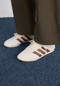 Hvide sneakers med brune striber, iført brune bukser på et blåt tæppe.