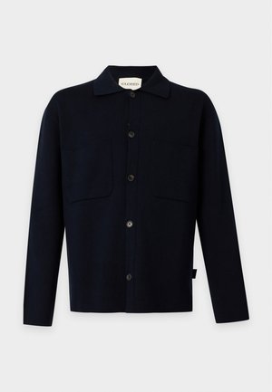Homme debout les mains dans les poches, portant une chemise noire boutonnée, un pantalon beige et des chaussures noires sur un fond blanc uni.