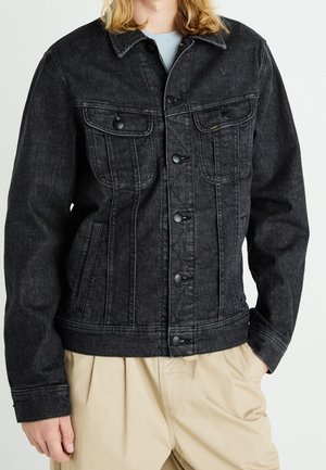 Person trägt eine schwarze Jeansjacke mit knöpfbaren Brusttaschen und beige Hose vor einem schlichten weißen Hintergrund.