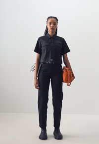 Black cropped button-up shirt with two front pockets, paired with black straight-leg pants and chunky black boots. Orange handbag accent.