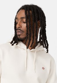 Cream-colored hoodie with a drawstring, soft texture, and a small branded label on the chest. Model with long, twisted hair facing slightly away.
