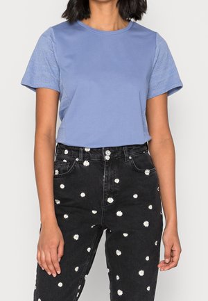 Woman wearing a light blue textured short-sleeve top paired with high-waisted black jeans featuring a white daisy print pattern.