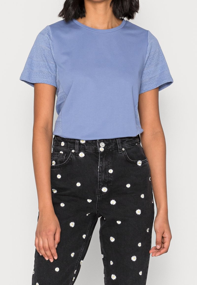 Femme portant un haut court texturé bleu clair associé à un jean noir taille haute avec un motif imprimé marguerites blanches.