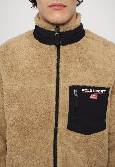 Tan fleece jacket with a high collar, black zippered chest pocket, and contrasting black neckline. Features a zipper closure and textured fabric.