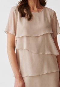 Vaalea beige kerrospusero, jossa on lyhyet hihat, teksturoidut pystysuorat raidat ja pyöreä pääntie. Pehmeä, kevyt kangas.