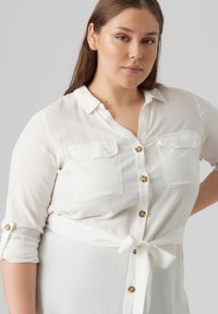 Camisa blanca de botones, hecha de una tela ligera, con dos bolsillos en el pecho, mangas remangadas y un acento de cintura atada.