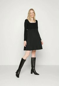 Mulher sorridente vestindo um vestido preto até ao joelho com mangas longas e botas de salto alto pretas até ao joelho, em pé contra um fundo claro e liso.