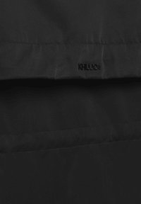 Črna tkanina z vodoravnimi šivi in majhnim črnim vezenim logotipom "KHUJO" blizu šiva.