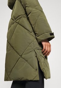 Abrigo acolchado color oliva con diseño acolchado, abertura lateral y cierre de broche en el dobladillo. Los puños tienen un detalle de velcro para un ajuste ajustable.