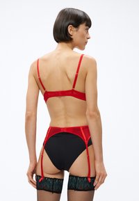 Červená saténová podprsenka s nastaviteľnými ramienkami, doplnená čiernymi nohavičkami s vysokým pásom a červeným podväzkovým pásom, s modrými pančuchami s čipkovaným vzorom.