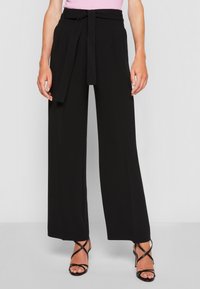 Pantalons larges noirs à taille haute avec une ceinture en tissu. Texture lisse, devant plissé, assortis avec des sandales à talons hauts noires à lanières.