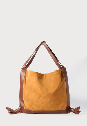 Sac fourre-tout en daim doux couleur moutarde avec des bords en cuir marron, des sangles pour épaule et des extrémités nouées de chaque côté, sur fond blanc.