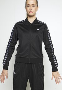 Svart träningsdräkt i glansigt tyg; zip-up-jacka med lila logotyp på ärmarna, matchande lösa byxor med elastisk midja.