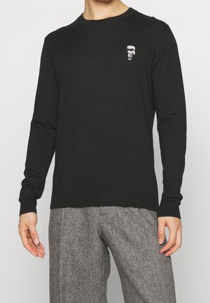 Mann trägt einen schwarzen Langarmpullover mit kleinem weißem Cartoon-Logo auf der Brust und graue Wollhosen.
