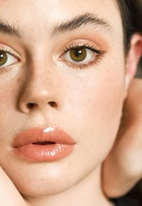 Nærbilde av et ansikt med glatt hud, middels ferskenfarget leppestift, markerte øyenbryn og varme hasselgrønne øyne. Diskret, naturlig sminke.