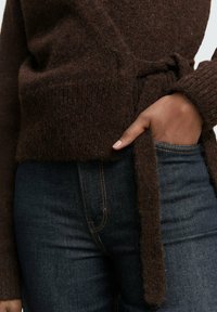 Bruin gebreid sweater met een strikceintuur, met een gestructureerde afwerking. Gepaarde met donkerblauwe denim jeans, die een aansluitende uitstraling geven.