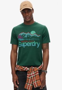 Grøn bomulds T-shirt med bjerg- og solgrafik i lilla og blå samt "Superdry" tekst. Båret med en ternet skjorte bundet om taljen.