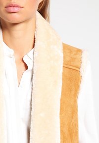 Ljusbrun mockaväst med en krämfärgad fuskpäls-kant, med en mjuk textur och en enkel design. Bäraren står framför en neutral bakgrund.