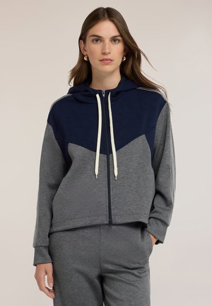 Vrouw draagt een tweekleurige marineblauwe en grijze hoodie met ritssluiting, witte trekkoorden en bijpassende grijze broek, staand tegen een effen achtergrond.