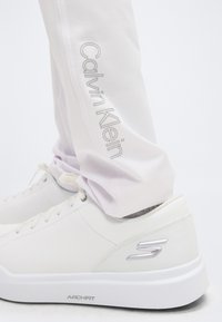 Κοντινό πλάνο λευκών αθλητικών παπουτσιών Skechers Arch Fit που φοριούνται με ανοιχτόχρωμο παντελόνι Calvin Klein, με εμφανή τα λογότυπα της μάρκας στο παπούτσι και το πόδι του παντελονιού.