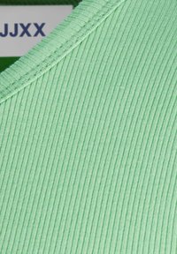 Närbild av ljusgrön ribbad tyg med synlig sömn och en del av en vit etikett som visar mörkblå text "JJXX" på grön bakgrund.
