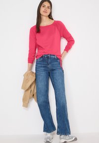 Femme en haut rose à manches longues, jean bleu et baskets argentées tenant une veste beige devant un fond blanc uni.