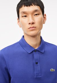 Camisa polo de un azul vibrante, confeccionada en algodón texturizado. Presenta un cierre de dos botones, mangas cortas y un pequeño logo verde en el pecho.