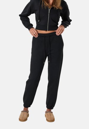 Kobieta ubrana w czarną, krótką bluzę na zamek z kapturem, czarne spodnie dresowe ze sznurkiem oraz beżowe kapcie wsuwane z ozdobnym wzorem.