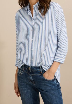 Kobieta ubrana w luźną, zapinaną na guziki białą koszulę z pionowymi niebieskimi paskami oraz ciemnoniebieskie dżinsy, z jedną ręką w kieszeni, na jednolitym tle.