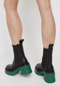 Botas de tobillo de cuero negro con paneles laterales elásticos y un tacón grueso verde, con punta redondeada y suela texturada.