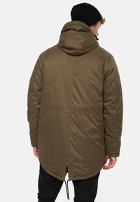Parka impermeabilă de culoare verde măsliniu, cu tiv cu șnur, glugă mare și detalii de cusătură. Beanie negru purtat, arătat din spate.