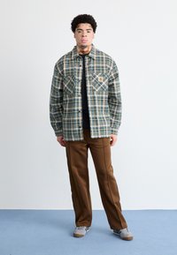 Stribet skjorte i grøn, blå og creme med to brystlommer. Sammen med brune bukser og grå sneakers på et lyseblåt gulv.