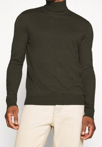 Khaki golf z wysokiej jakości dzianiny, z długimi rękawami oraz ściągaczami u rękawów i na dole, w zestawie z jasnobeżowymi spodniami.