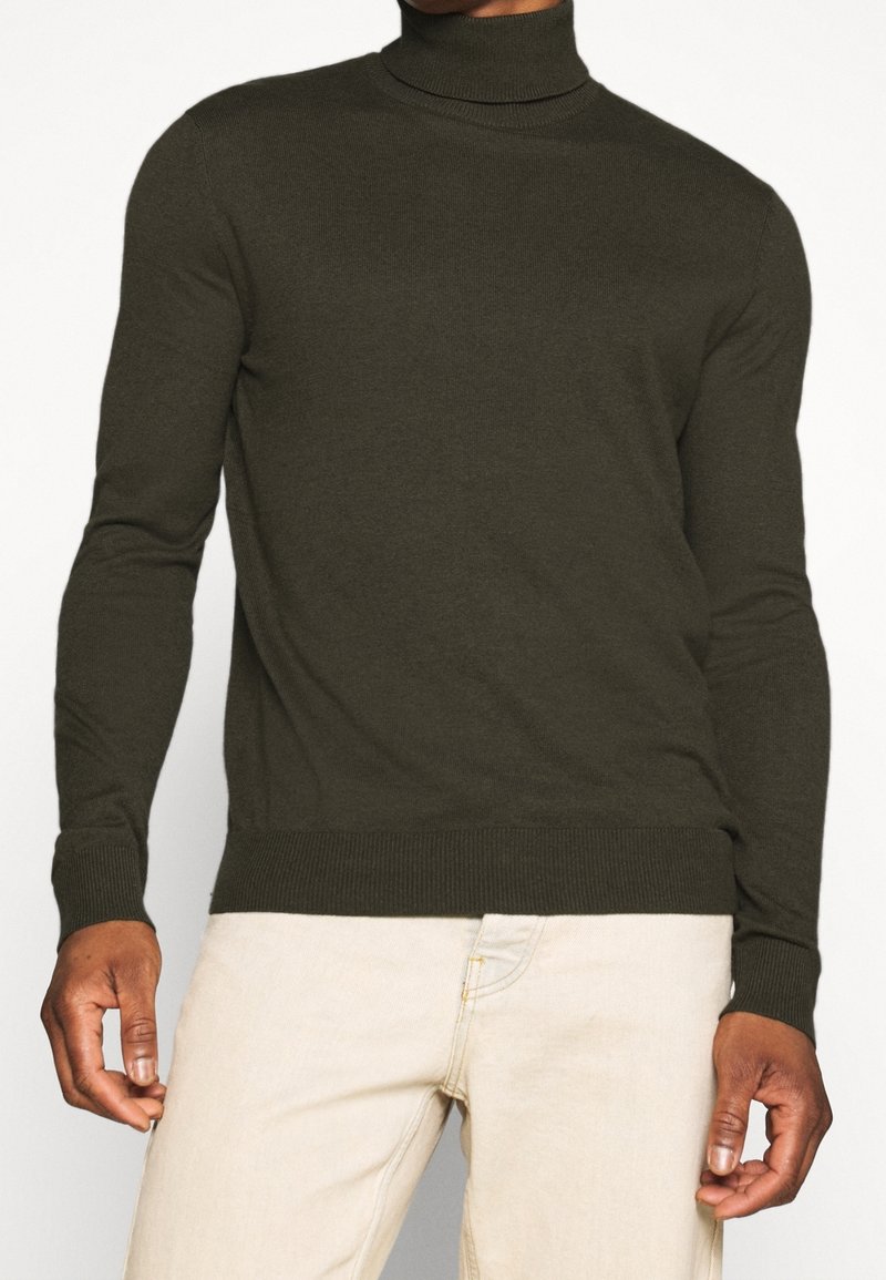 Khaki golf z wysokiej jakości dzianiny, z długimi rękawami oraz ściągaczami u rękawów i na dole, w zestawie z jasnobeżowymi spodniami.